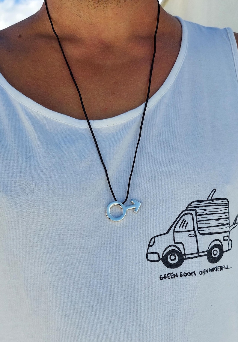 Puede incluir: Un collar de cord&oacute;n negro con un colgante plateado en forma de s&iacute;mbolo masculino. El collar lo lleva una persona que lleva una camiseta blanca de tirantes con una ilustraci&oacute;n en blanco y negro de una furgoneta con el texto "GREEN ROOM open waterfall..." en la parte delantera.