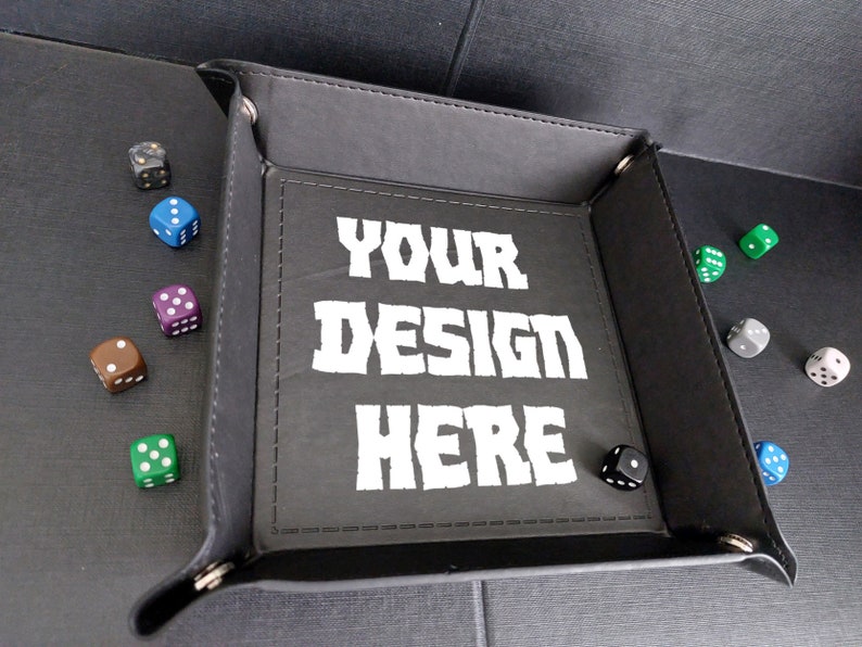 Pu&ograve; includere: Vassoio da gioco in feltro nero con una scritta bianca che dice "YOUR DESIGN HERE". Il vassoio &egrave; circondato da dadi di vari colori.