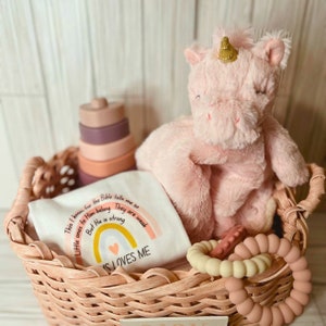 Puede incluir: Una cesta de mimbre llena de regalos para bebés, que incluyen un juguete de peluche de unicornio rosa, un body blanco con un arcoíris y el texto "This I know for the Bible tells me so Little ones to Him belong They are weak But He is strong IS LOVES ME", y un juego de anillos de dentición de silicona beige y marrón.