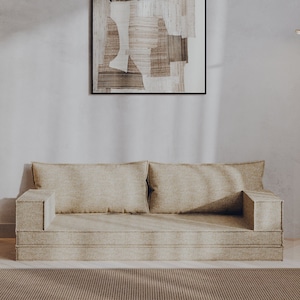 Puede incluir: Un sofá beige con una alfombra marrón claro delante. Una lámpara de pie con una base dorada y una pantalla blanca está en la esquina de la habitación. Un cuadro abstracto enmarcado cuelga de la pared detrás del sofá.