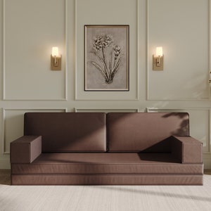 May include: A brown futon sofa with a light brown rug in front of it. The sofa is in a room with white walls and gold accents. There are two wall sconces on either side of a framed print of a floral design. There is a small table with a vase of white flowers on it to the left of the sofa and a large potted plant to the right.