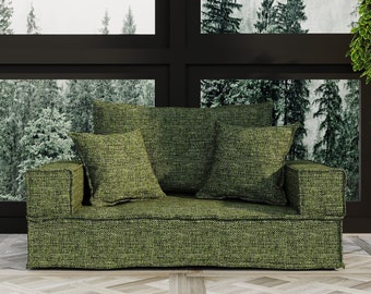 May include: A green fabric sofa with two pillows sits in front of a large window with a view of a snowy forest. The sofa is in a room with a wood floor.