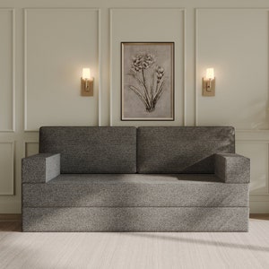 Op de afbeelding: Een grijze stoffen bank met twee kussens staat in een kamer met witte muren en een beige kleed. Aan weerszijden van een ingelijst drukwerk met een bloemenmotief bevinden zich twee gouden wandlampen met witte glazen kapjes.