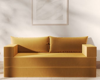 May include: A mustard yellow sofa with two cushions sits in a minimalist living room with a white wall and a framed picture above it. A small white side table with a glass and a book sits to the left of the sofa.