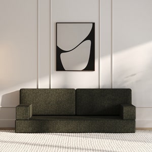 Op de afbeelding: Een donkergroene bank met een wit tapijt ervoor. Er is een zwarte staande lamp met twee witte bolvormige lampen, een ingelijst abstract kunstwerk aan de muur en een kleine houten bijzettafel met een vaas met groen op.