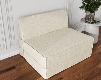 May include: A white fabric futon sofa with a simple design. The sofa is folded into a sitting position and is sitting on a wooden floor.