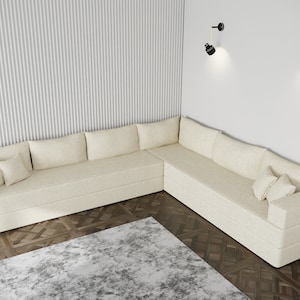 Può includere: Un grande divano angolare di colore bianco crema con più cuscini si trova in una stanza con una parete bianca e un pavimento in legno. Due applique da parete nere con dettagli dorati sono montate sulla parete.