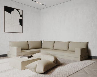 May include: A beige sectional sofa with a low profile and a modern design. The sofa is placed on a light beige rug in a room with white walls and a white ceiling. There are two black and white abstract paintings on the wall behind the sofa.