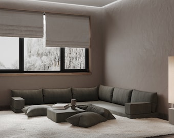 Può includere: Un grande divano angolare a forma di L di colore grigio con più cuscini è posizionato su un tappeto bianco a pelo lungo in una stanza con una finestra che si affaccia su un bosco innevato. La stanza ha una parete grigio chiaro e un pavimento in legno.