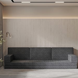 Op de afbeelding: Een moderne woonkamer met een donkergrijze bank met patroon en een bijpassend bijzettafeltje. De kamer heeft een neutraal kleurenpalet met een lichtgekleurde muur en vloerkleed. Een plant en een lamp zijn ook zichtbaar.