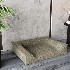 May include: A beige dog bed with a raised edge and a textured fabric. The bed is sitting on a wooden floor in front of a concrete wall.