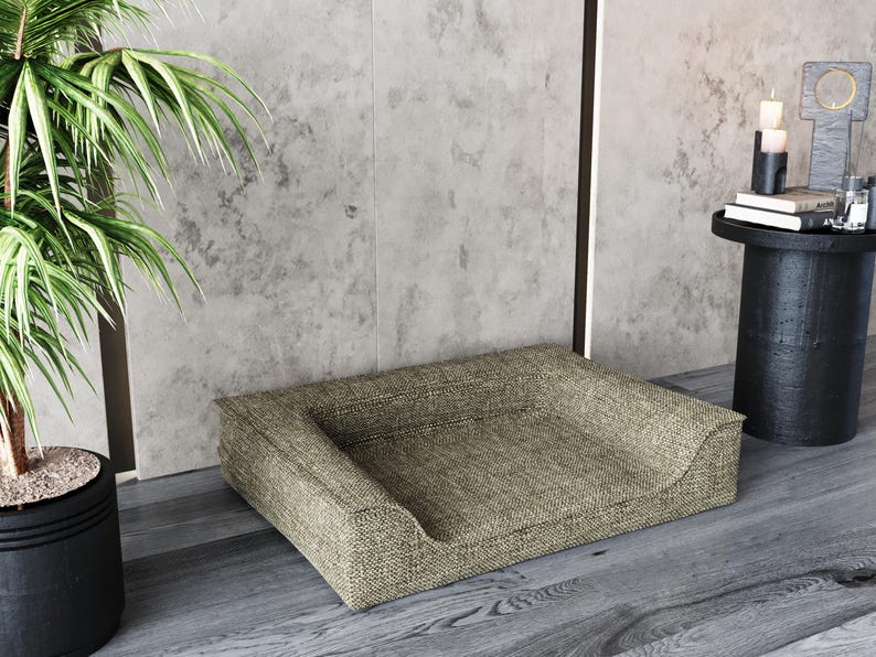 May include: A beige dog bed with a raised edge and a textured fabric. The bed is sitting on a wooden floor in front of a concrete wall.