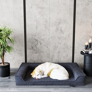 May include: A white and brown dog sleeping on a dark blue dog bed with a raised edge. The bed is on a gray concrete floor with a black plant pot and a black side table with candles and a glass vase.