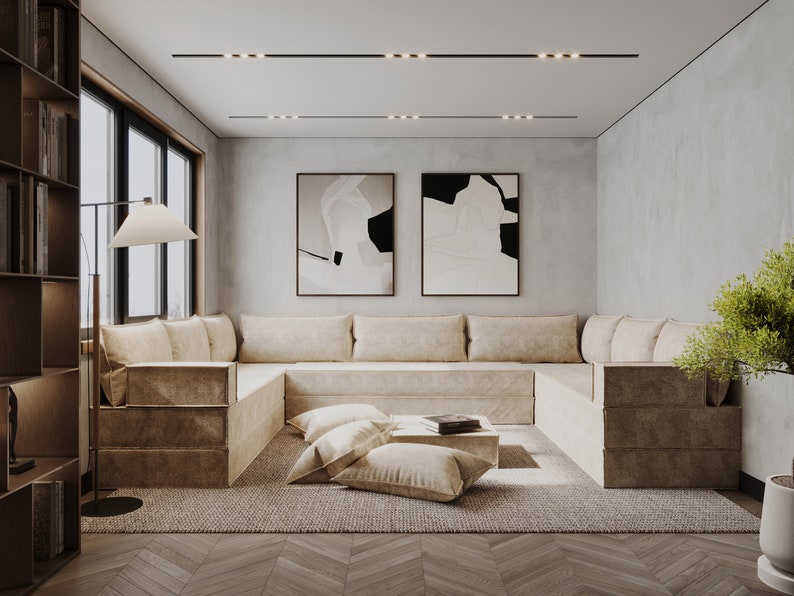 May include: A large, light brown sectional sofa with multiple cushions sits in a modern living room with a light gray wall, a wood floor, and two abstract black and white paintings.