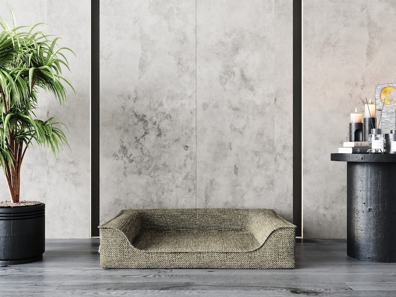May include: A light brown dog bed with a raised edge and a soft, textured fabric. The bed is sitting on a gray concrete floor in front of a gray wall.