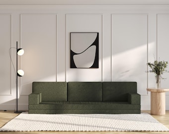 May include: A dark green sofa with three cushions sits in a white room with a black floor lamp and a small wooden side table with a vase of greenery. A black framed abstract art print hangs on the wall.