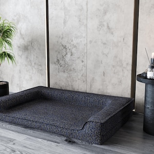 May include: A dark blue dog bed with a raised rim and a soft, textured surface. The bed is sitting on a wooden floor in front of a gray wall.