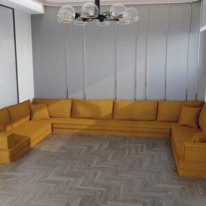 May include: A large, mustard yellow sectional sofa with multiple cushions and a low profile. The sofa is in a modern living room with a wood floor and a chandelier.