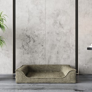 Peut inclure: Un panier pour chien beige avec une texture tiss&eacute;e, plac&eacute; devant un mur gris. Le lit a un profil bas avec des c&ocirc;t&eacute;s sur&eacute;lev&eacute;s. Un pot de plante et une petite table avec des bougies et des ornements sont &eacute;galement visibles.