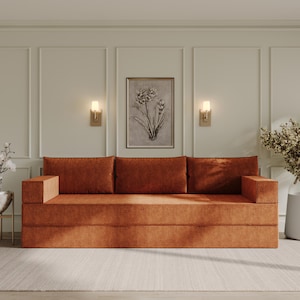 Puede incluir: Un sofá de terciopelo naranja quemado con tres cojines y una alfombra marrón claro debajo. El sofá está en una habitación con paredes blancas y apliques de pared dorados.