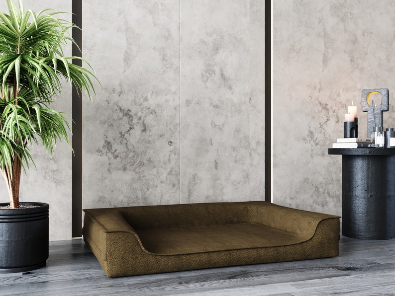 May include: A brown dog bed with a raised edge and a slightly indented center. The bed is sitting on a light wood floor in front of a concrete wall.