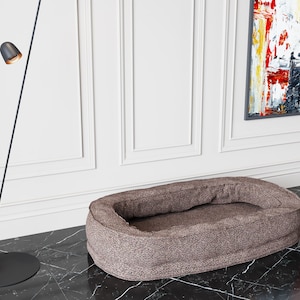 Puede incluir: Una cama para perro de forma ovalada de color marrón con un tejido suave y de felpa. La cama está sobre un suelo de baldosas blancas y negras.