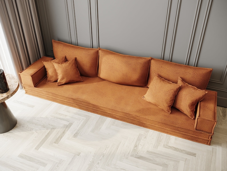 May include: A long, low, orange-brown sofa with a tufted back and armrests. The sofa is covered in a soft, textured fabric and has several matching throw pillows. The sofa is sitting on a light wood floor with a herringbone pattern.