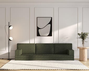 May include: A dark green sofa with three cushions sits in a room with white walls and a black floor lamp. A white rug is in front of the sofa. A small wooden side table with a vase of greenery sits to the right of the sofa. A black framed print with an abstract design hangs on the wall.