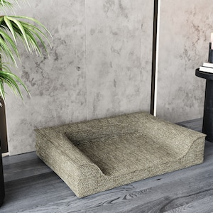 Peut inclure: Un lit pour animaux de compagnie rectangulaire avec un devant bas et incurv&eacute; et un tissu tiss&eacute; beige. Le lit est plac&eacute; sur un sol en bois, pr&egrave;s d'une plante en pot et d'une petite table d'appoint noire avec des bougies et des livres.
