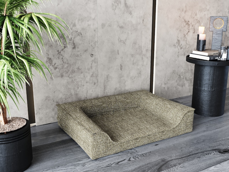 Peut inclure: Un lit pour animaux de compagnie rectangulaire avec un devant bas et incurv&eacute; et un tissu tiss&eacute; beige. Le lit est plac&eacute; sur un sol en bois, pr&egrave;s d'une plante en pot et d'une petite table d'appoint noire avec des bougies et des livres.