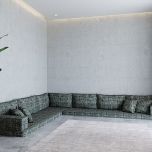 Op de afbeelding: Een grote, donkergroene hoekbank met meerdere kussens staat in een moderne woonkamer met een betonnen muur en een lichtkleurig tapijt.