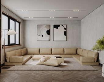 May include: A large, light brown sectional sofa with multiple cushions and pillows sits on a beige rug in a modern living room. The room has a concrete wall with two abstract paintings and a small potted plant in the corner.