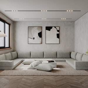 May include: A modern living room with a large, light gray sectional sofa, a low coffee table, and a rug. The room has a minimalist design with a concrete wall and two abstract paintings.