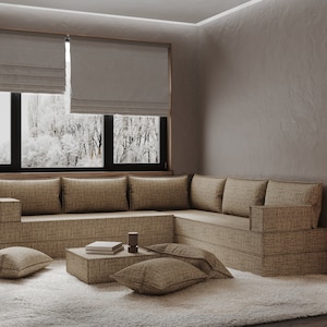 May include: A large, light brown sectional sofa with a low, square coffee table in front of it. The sofa is made of a textured fabric and has a variety of cushions. The coffee table is made of the same fabric as the sofa and has a few books stacked on top of it. The room is decorated in a minimalist style with a white rug and a large window with blinds.