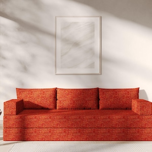 May include: A red fabric sofa with three cushions in a minimalist living room setting. The sofa is in front of a white wall with a framed picture above it.