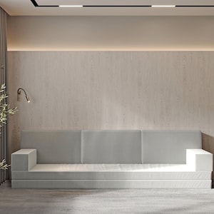 Puede incluir: Un sofá seccional gris claro con una pared de madera detrás. El sofá tiene un diseño simple y está ubicado en una sala de estar moderna.