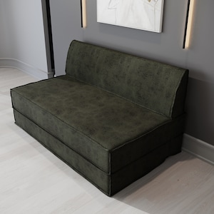 May include: A dark green, two-seater sofa with a textured surface. The sofa has a low back and a rectangular shape. The stitching is visible along the edges. The sofa is placed against a gray wall.