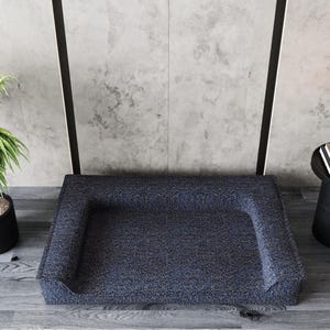 Peut inclure: Un lit pour animaux de compagnie bleu marine avec un motif mouchet&eacute;. Le lit a des c&ocirc;t&eacute;s sur&eacute;lev&eacute;s et est plac&eacute; sur un sol en bois. Une plante en pot et une petite table d'appoint noire avec des bougies et des livres sont en arri&egrave;re-plan.