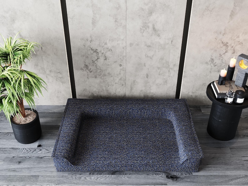 Peut inclure: Un lit pour animaux de compagnie bleu marine avec un motif mouchet&eacute;. Le lit a des c&ocirc;t&eacute;s sur&eacute;lev&eacute;s et est plac&eacute; sur un sol en bois. Une plante en pot et une petite table d'appoint noire avec des bougies et des livres sont en arri&egrave;re-plan.