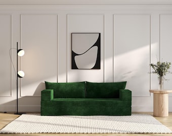 May include: A green velvet sofa with two pillows sits in a room with white walls and a white rug. A black floor lamp with two white globes stands to the left of the sofa. A small wooden side table with a vase of flowers sits to the right of the sofa. A black framed print with a white abstract design hangs on the wall behind the sofa.