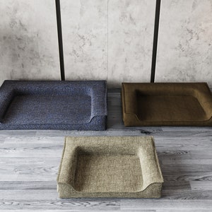 May include: Three dog beds in different colors and textures. The beds are rectangular with a raised edge and a dip in the center. The beds are on a wooden floor.