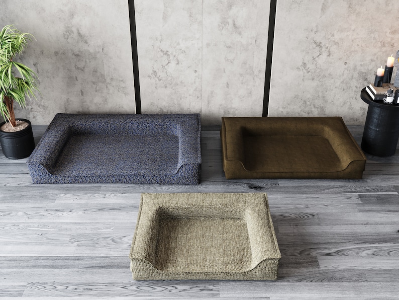 May include: Three dog beds in different colors and textures. The beds are rectangular with a raised edge and a dip in the center. The beds are on a wooden floor.