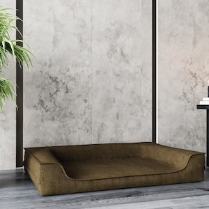 May include: A brown dog bed with a raised edge and a slightly indented center. The bed is sitting on a light wood floor in front of a concrete wall.
