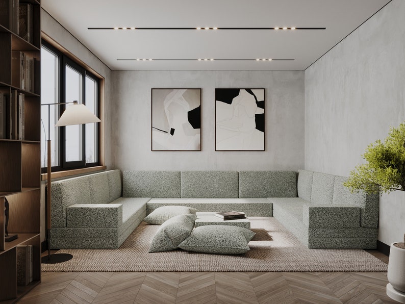 May include: A modern living room with a large, light green sectional sofa, a coffee table with a book on it, and two abstract paintings on the wall. The room has a light wood floor and a rug.