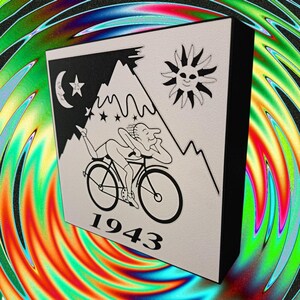 Può includere: Un'opera d'arte quadrata in bianco e nero raffigura una persona sdraiata su una bicicletta, con montagne, un sole, una luna e stelle. L'anno "1943" è stampato sotto. Ha bordi neri.