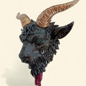 May include: A detailed sculpture of a goat's head with bronze-colored horns, a black pentagram on its forehead, and a red and purple rope-like detail. The goat's face is dark with visible teeth and a textured surface.