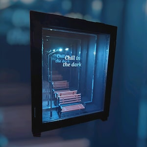 Może przedstawiać: Miniaturowa diorama w czarnej ramie, przedstawiająca scenę uliczną z ławkami i latarniami. Tekst "Chill in the dark" wyświetlany jest w neonowym, niebieskim świetle, tworząc nastrojową atmosferę. Ławki są w kolorze czerwonobrązowym.