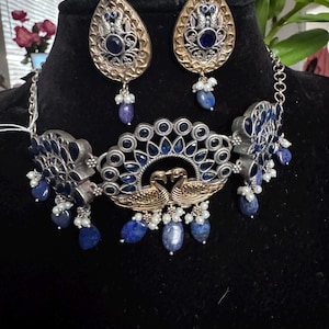 May include: A silver and gold necklace with a peacock design and blue and white stones. The necklace is paired with matching earrings.