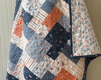 May include: A patchwork quilt with a blue, white, and orange color scheme. The quilt is made of various fabrics with different patterns, including floral, striped, and geometric designs. The quilt is folded and displayed on a wooden stand.