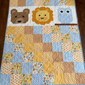 May include: A handmade baby quilt featuring a patchwork design with squares of blue, yellow, and brown fabrics. The quilt showcases appliqued animal faces: a bear, a lion, and an owl. The quilt is bordered with a light blue fabric.